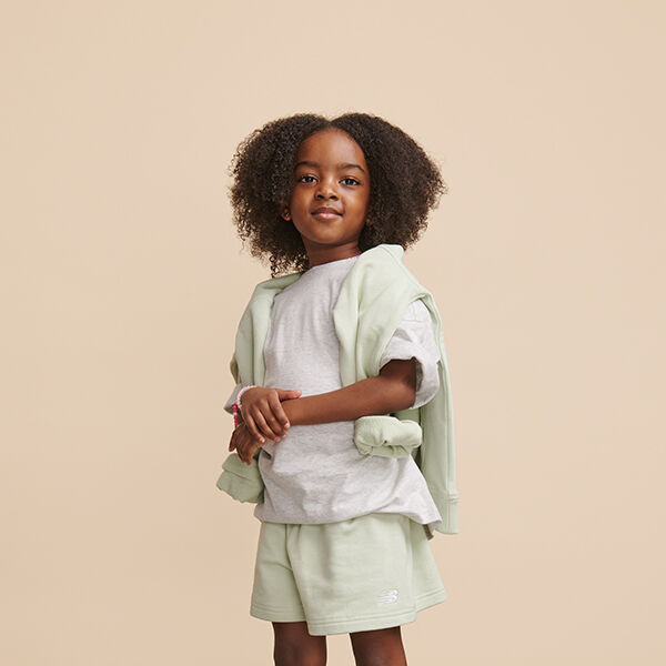 Young girl wearing light green New Balance sweatshirt over white shirt and light green shorts posed against beige background in studio environment 