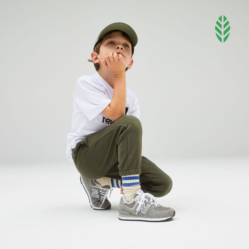 Boy wearing grey 574 Core shoes with white shirt, green hat and green sweatpants kneeling against a grey studio background with a green leaf icon in the upper right corner