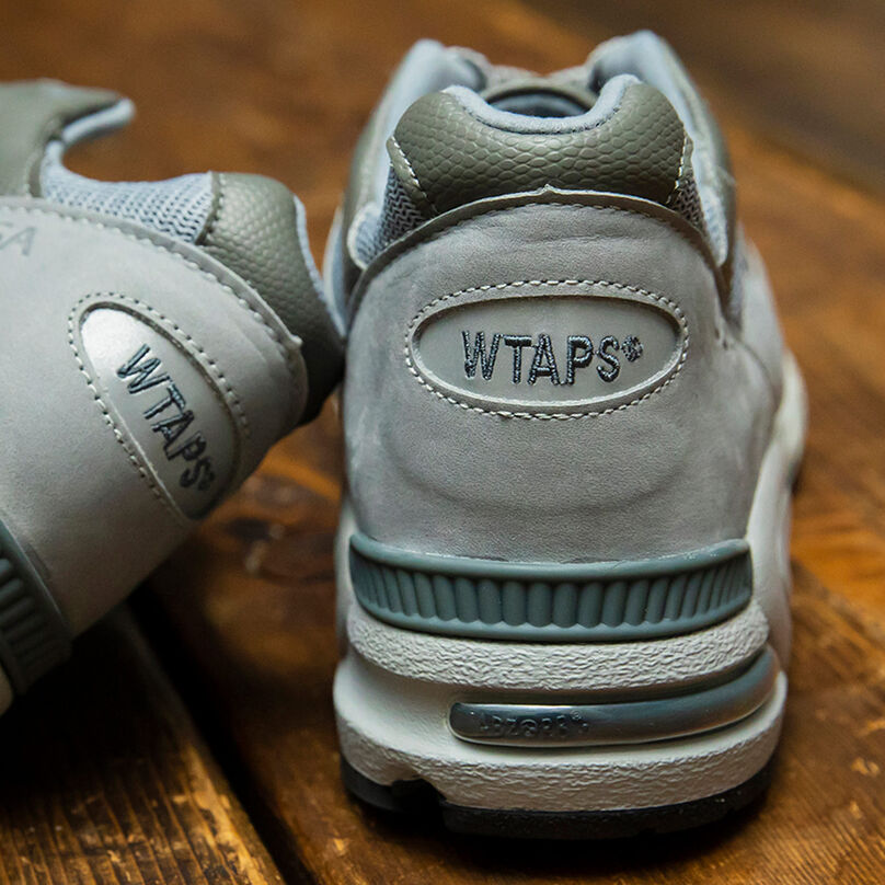 Close-up detail shot of the heels of a pair of grey NB x WTAPS 990v2 sneakers sitting on rustic wood flooring