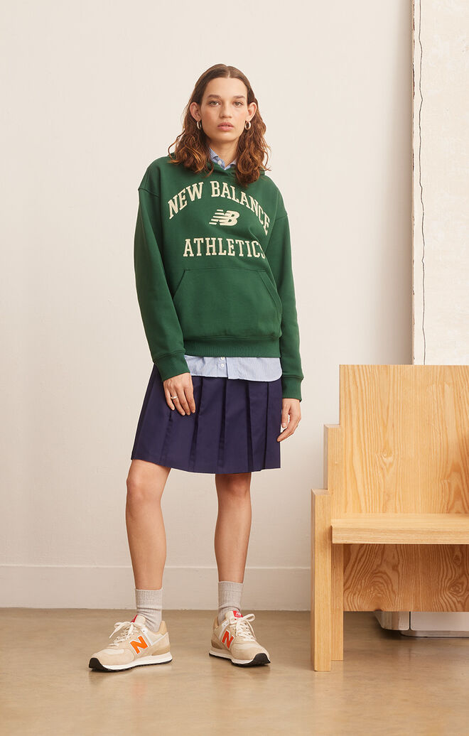 A young woman wearing a green NB Essentials hoodie, navy-blue pleated skirt, and NB 574 sneakers