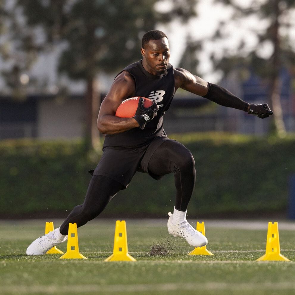Athlete wearing black training apparel runs a football agility drill on a grass field, holding a football while stepping between yellow cones.