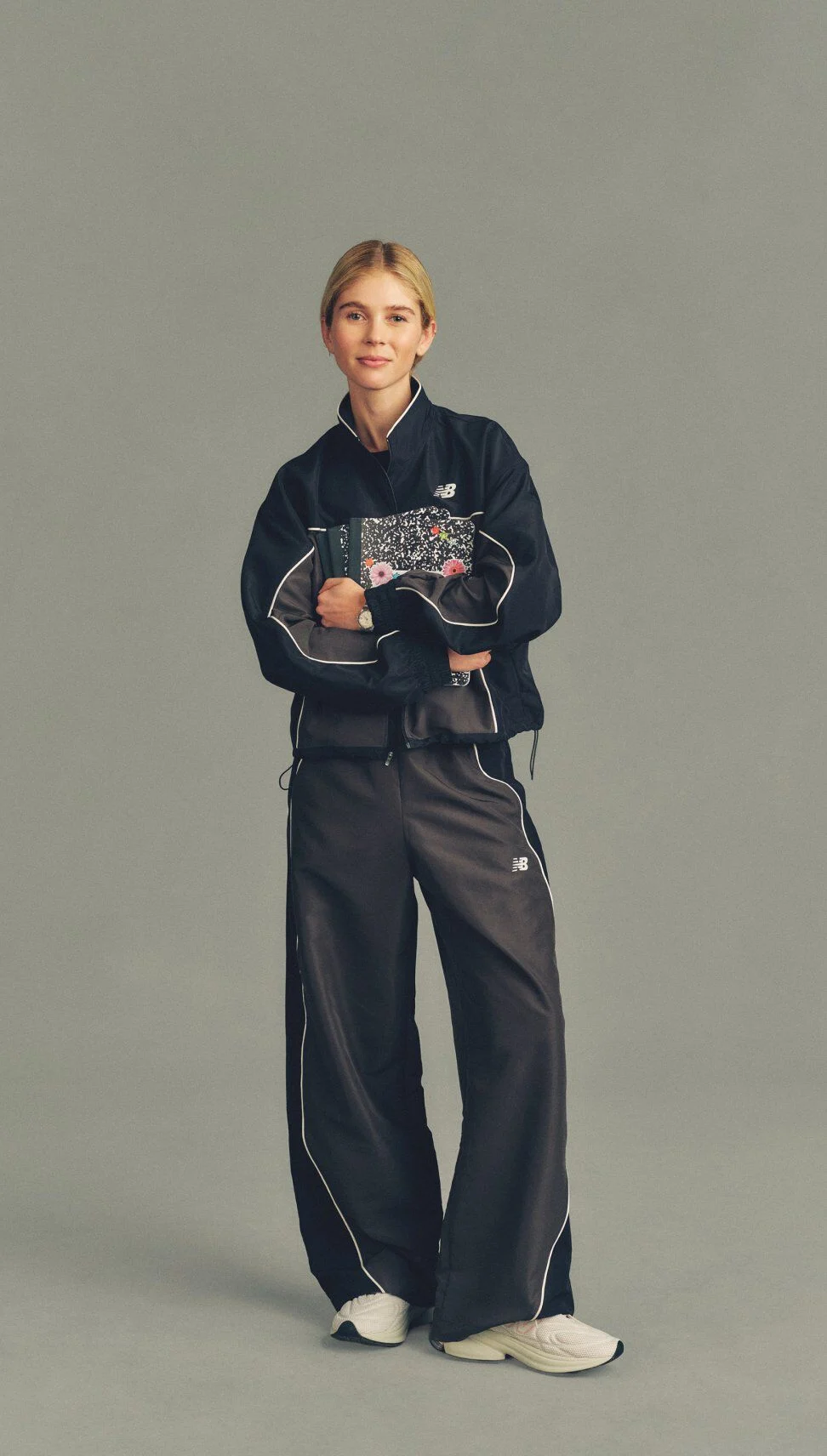 Full-body studio image of a person in a black New Balance tracksuit holding a patterned notebook against a neutral gray background.
