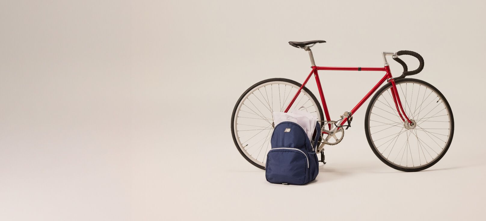 A red bicycle next to a blue New Balance backpack in front of a beige background.