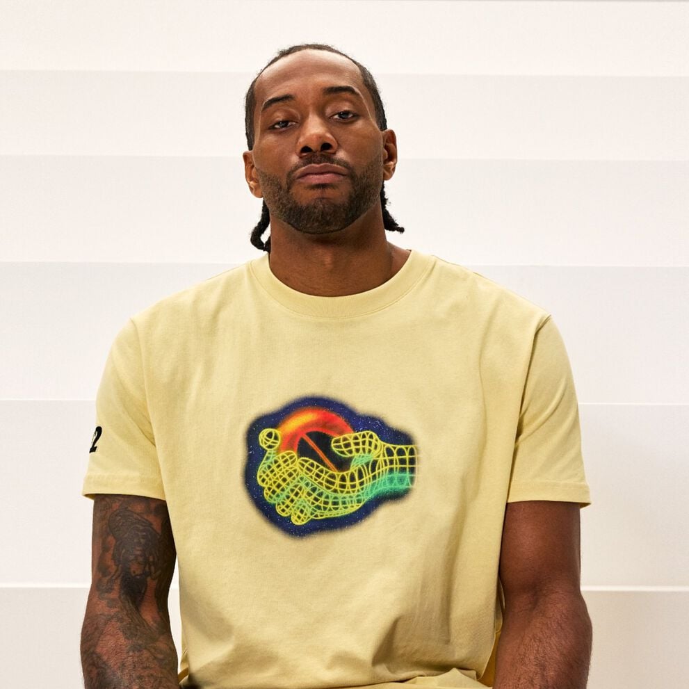 A person sits against a light, horizontally striped background wearing a pale yellow T‑shirt featuring a colorful graphic of two stylized, wireframe hands reaching toward each other around a glowing sphere.