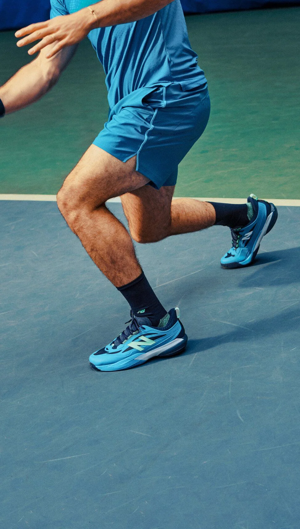 Tennis player lunges across an indoor court wearing bright blue New Balance tennis shoes during active play.