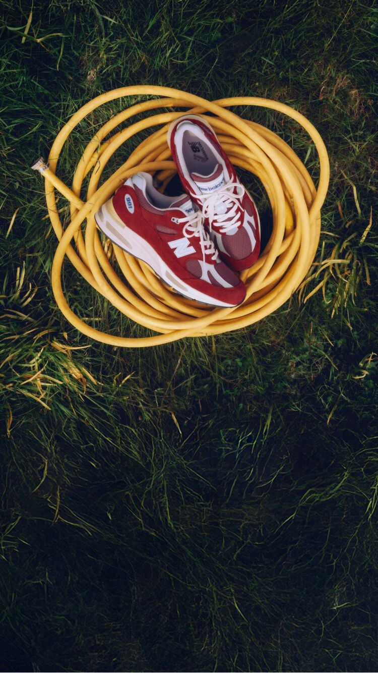 A pair of New Balance Made in UK sneakers sitting on a yellow coiled hose on top of a lush grass lawn.