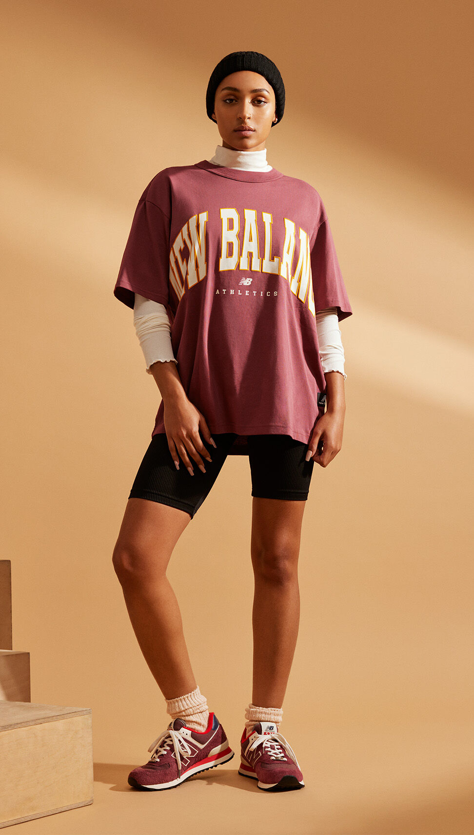 A young woman wearing a dark-red NB Uni-ssentials tee with black short tights and dark-red NB 574 sneakers in front of a burnt orange studio backdrop