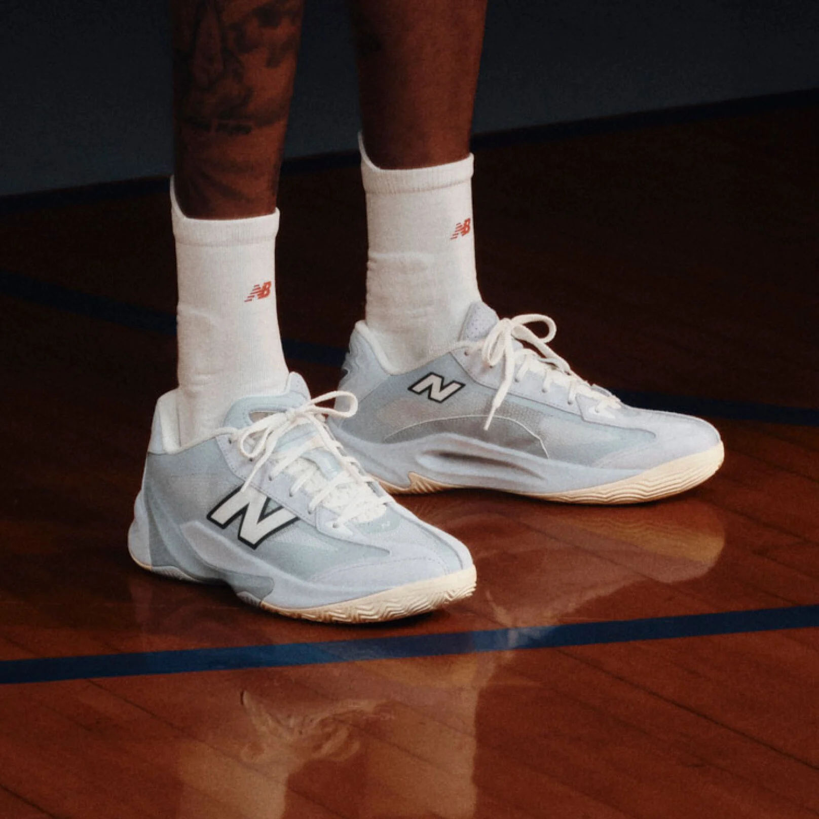 Light blue and white New Balance basketball shoes worn with white crew socks on a polished indoor basketball court.
