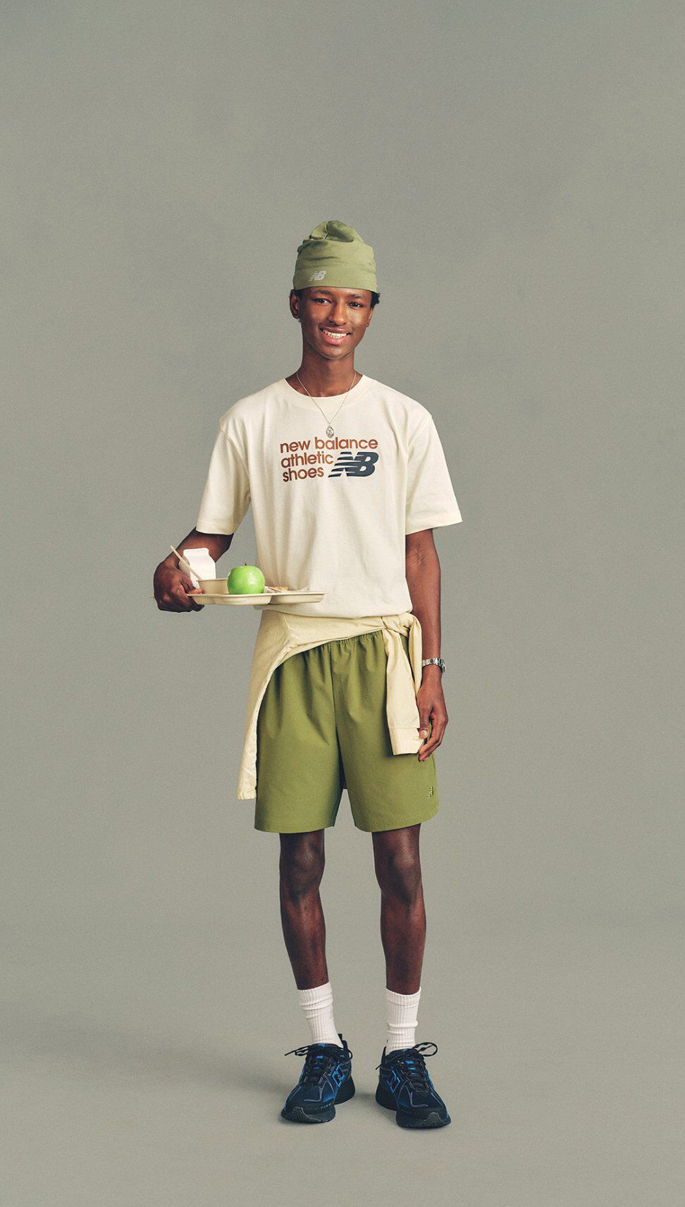 The person is wearing a white T-shirt with printed text reading &ldquo;new balance athletic shoes&rdquo; alongside the New Balance logo, paired with olive-green athletic shorts.