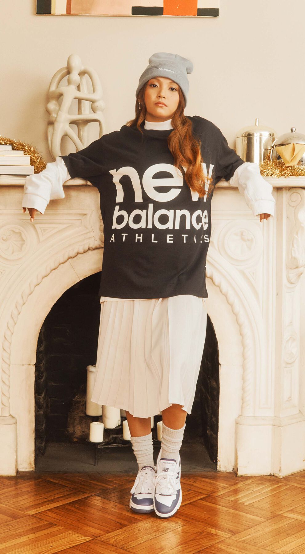 Female model wearing cold weather New Balance lifestyle clothing and shoes posed against a decorative fireplace
