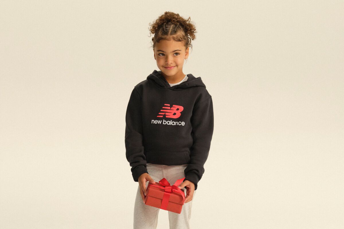 Child wearing a black New Balance hoodie and gray pants holding a red gift box against a light studio backdrop. 