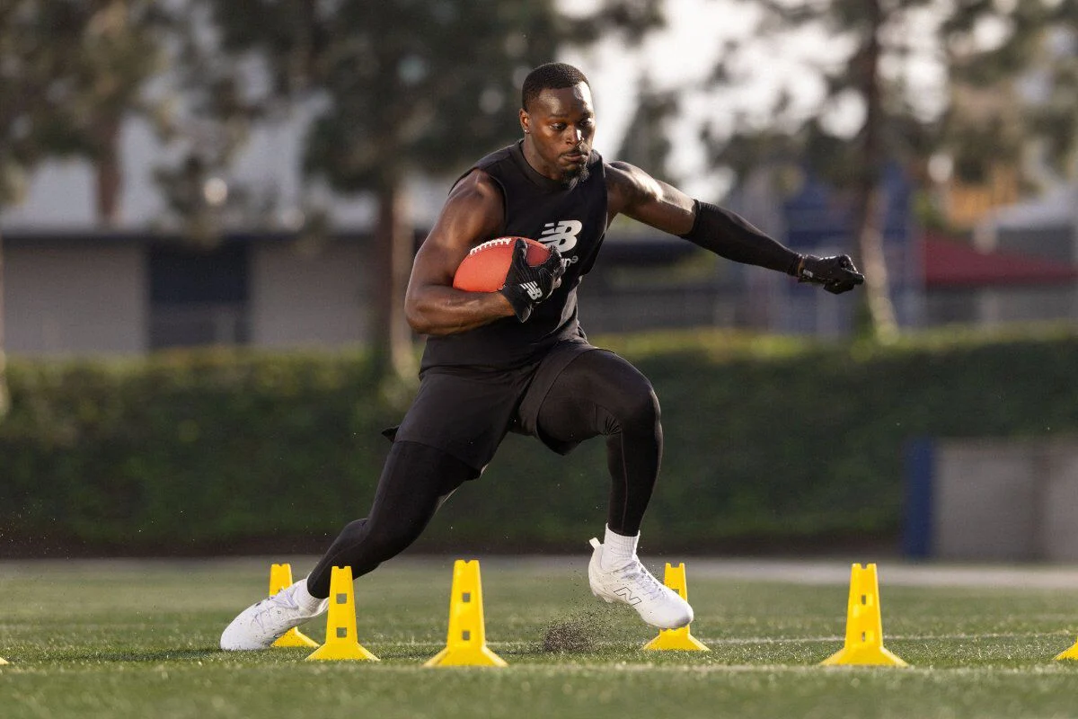 Athlete wearing black training apparel runs a football agility drill on a grass field, holding a football while stepping between yellow cones.