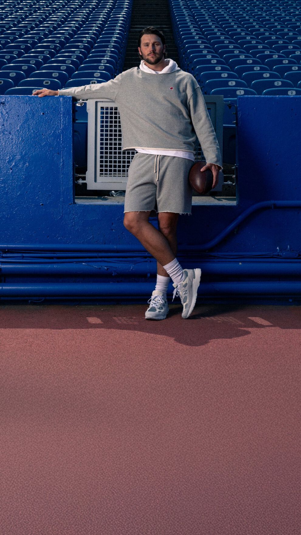 Young male athlete in a grey hoodie leaning on a stadium fence.