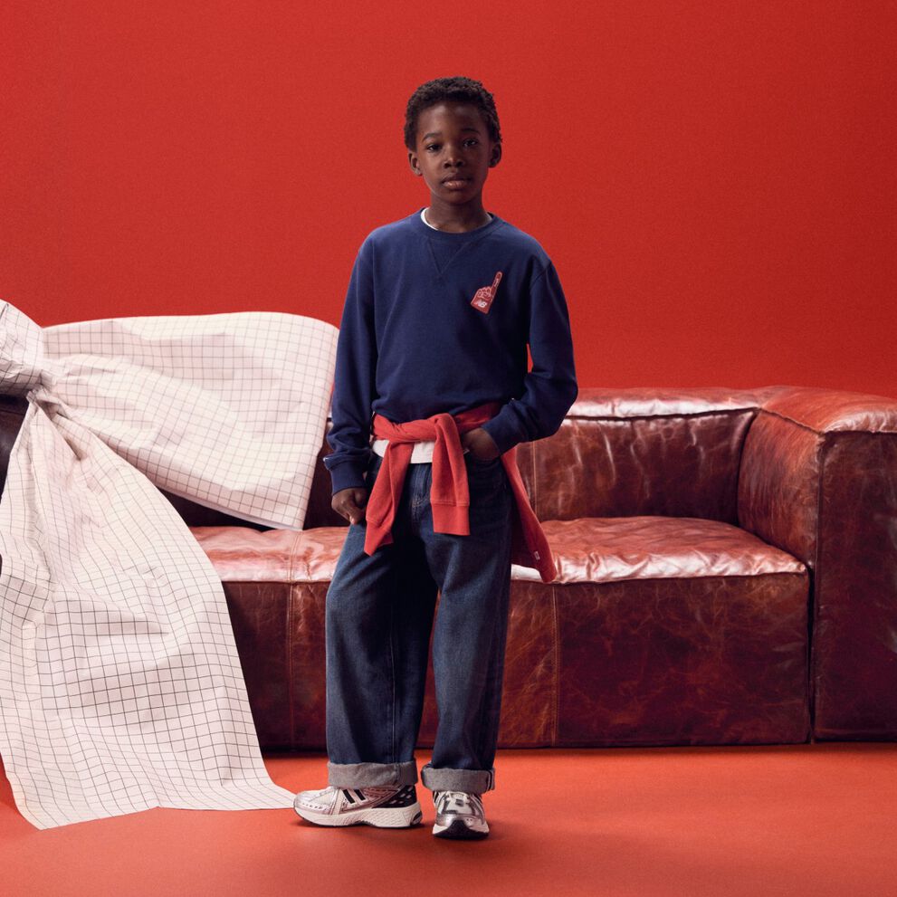Niño con una camisa de manga larga azul marino frente a un sofá rojo.
