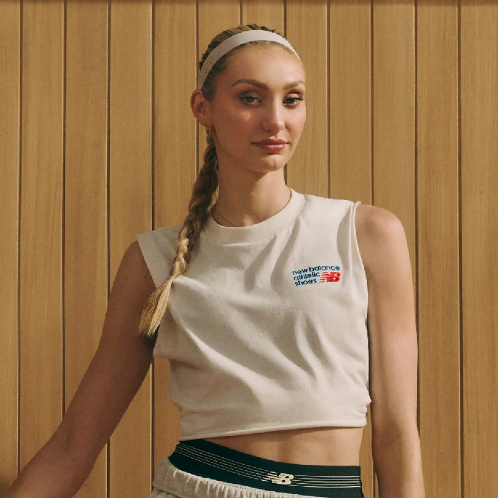 A person wearing a cropped athletic top and sport shorts stands in front of a wooden-paneled background, with a long braid over one shoulder.
