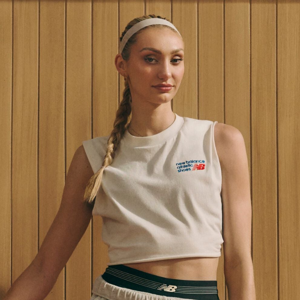 A person wearing a cropped athletic top and sport shorts stands in front of a wooden-paneled background, with a long braid over one shoulder.