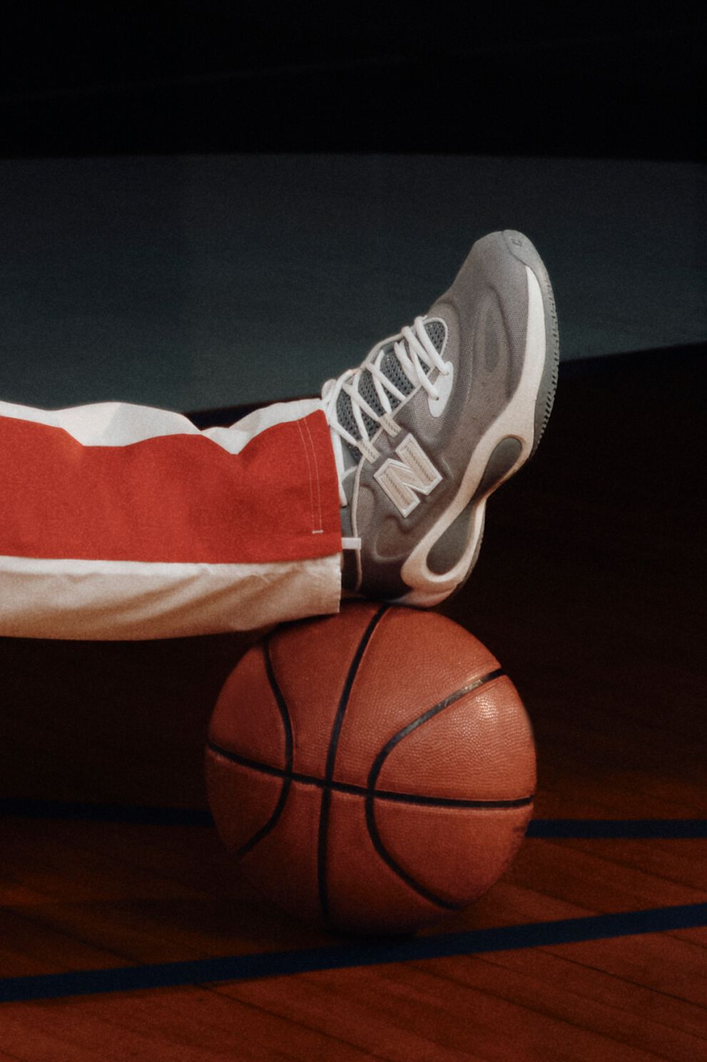 Gray New Balance basketball sneaker resting on a basketball, paired with white and red athletic pants on a dark court.