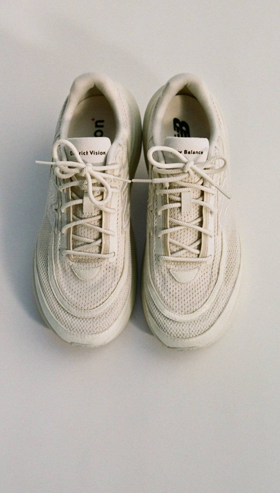 Top‑down view of a pair of off‑white New Balance athletic shoes placed side by side on a light background, showing mesh uppers, tonal laces, layered stitching, and visible New Balance and District Vision branding on the insoles and tongue