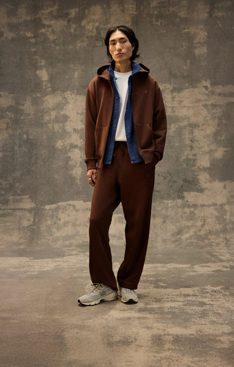 Person standing indoors against a textured backdrop, wearing a brown tracksuit layered over a blue inner piece and sneakers. 