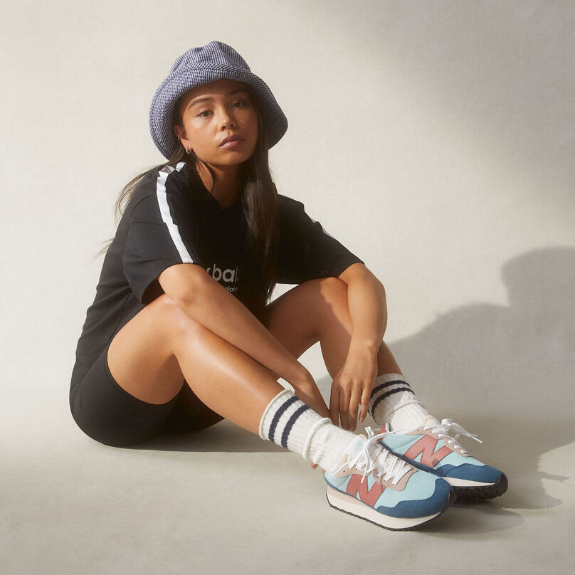 Girl wearing high socks with NB 237 shoes in blue, teal and salmon posed sitting in grey studio environment with shadow effects