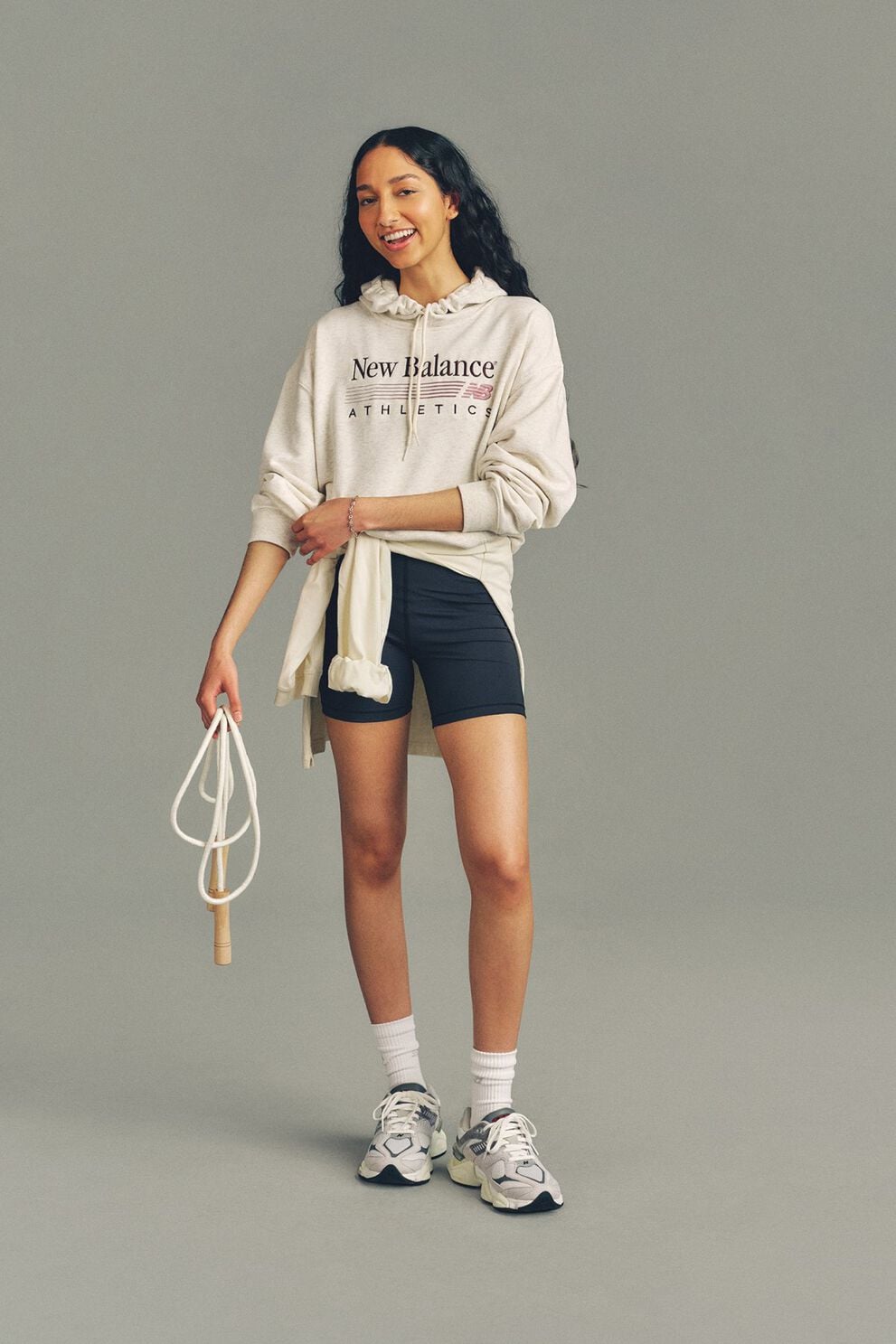 A woman wearing a New Balance hoodie and shorts standing in front of a grey studio background.