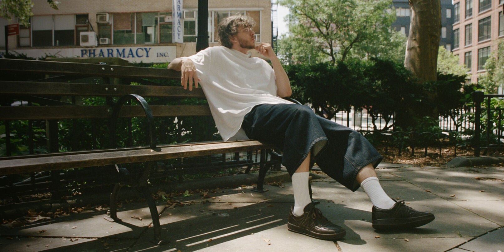Jack Harlow sitting outside on a park bench wearing dark brown New Balance sneakers.