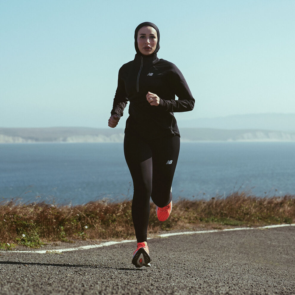 Ayah El Hafez running on paved road along shoreline wearing black New Balance jacket and tights with orange shoes 
