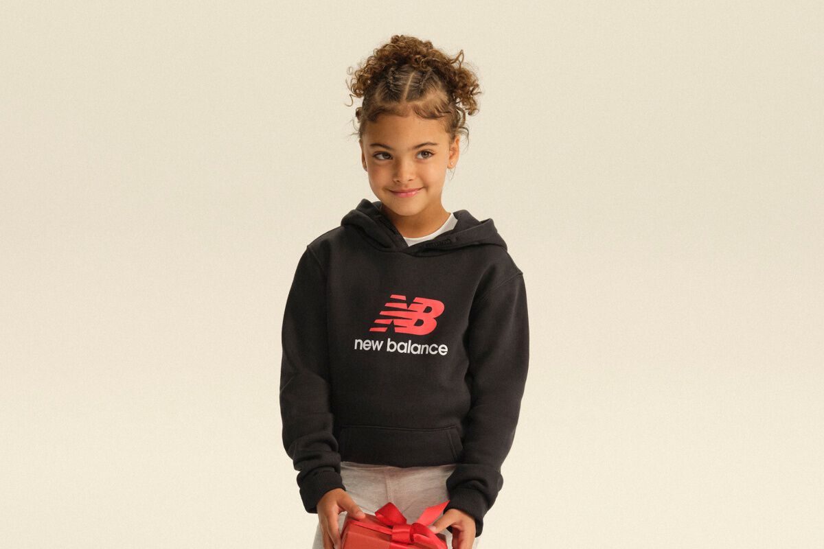 Child wearing a black New Balance hoodie and gray sweatpants while holding a red gift box in a clean studio setting.