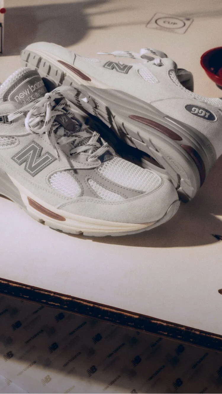 Un par de tenis Made in UK de New Balance sobre un banco de trabajo blanco, rodeado de herramientas para la fabricaci&oacute;n de calzado y otros equipos textiles.