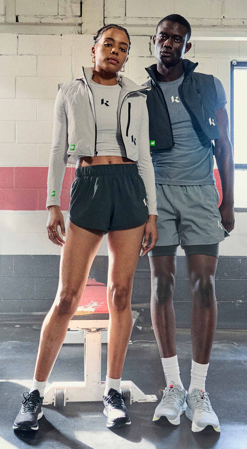 Man and woman wearing Klutch Athletics by New Balance, the man in a dark grey shirt, vest and shorts and the woman in a light grey shirt and vest with dark grey shorts posing in front of gym equipment 