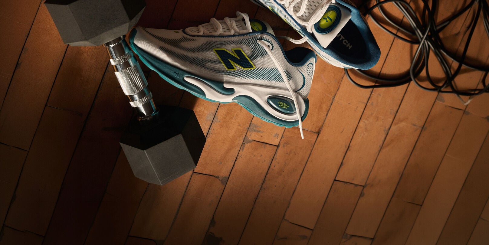 Close-up of New Balance training shoes placed beside a dumbbell and workout equipment on a hardwood gym floor.