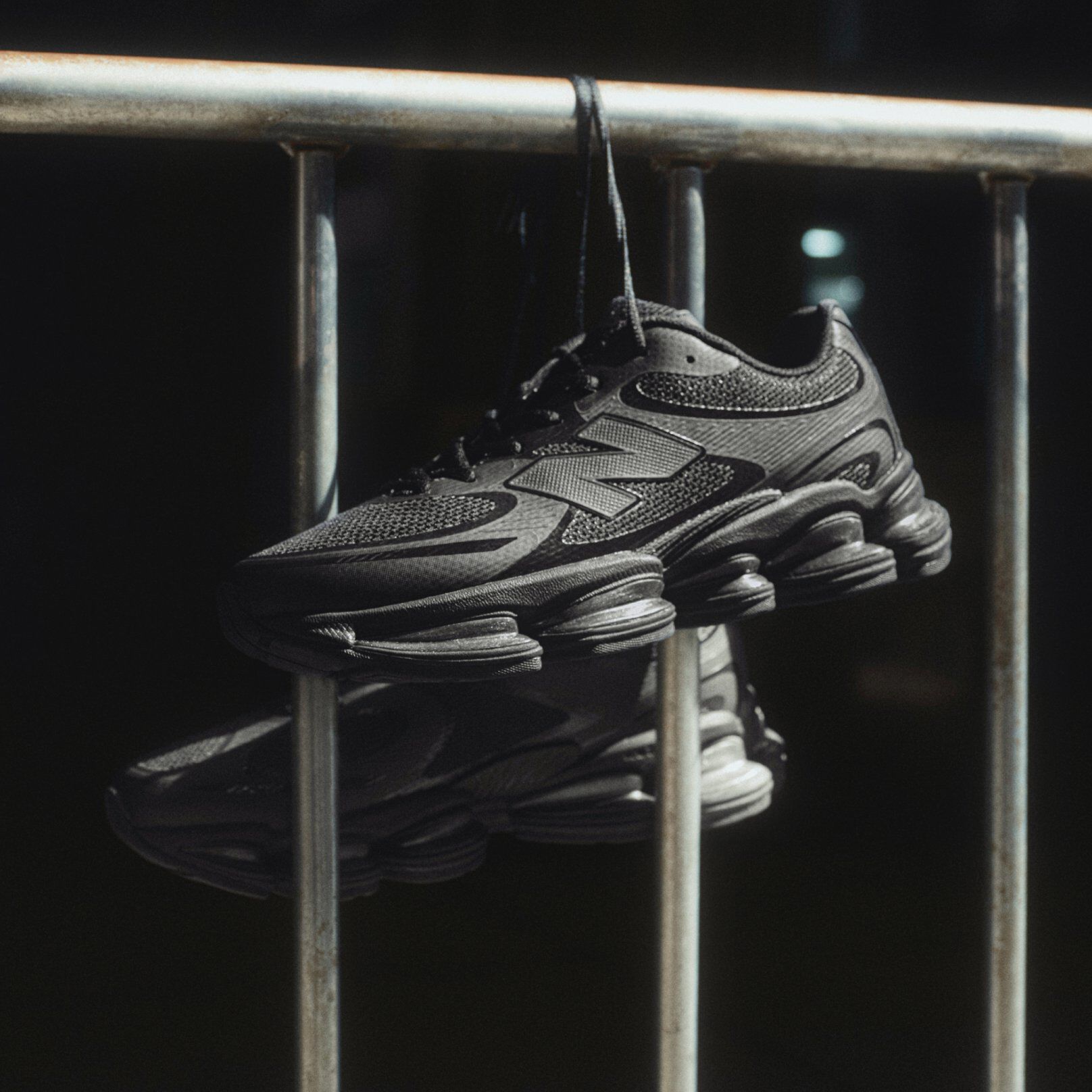 Black New Balance sneakers hanging by laces over a metal railing in dramatic lighting. 