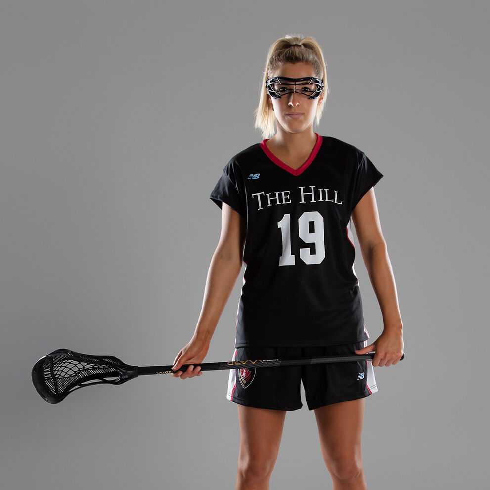 Female lacrosse player dressed in full gear posing in front of a grey studio background 