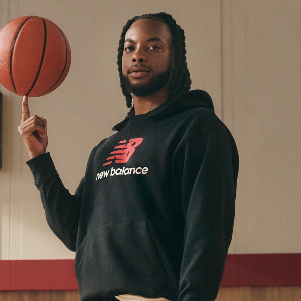 A person wearing a black New Balance hoodie with a red logo spins a basketball on their fingertip while standing indoors against a light-colored wall with red trim.