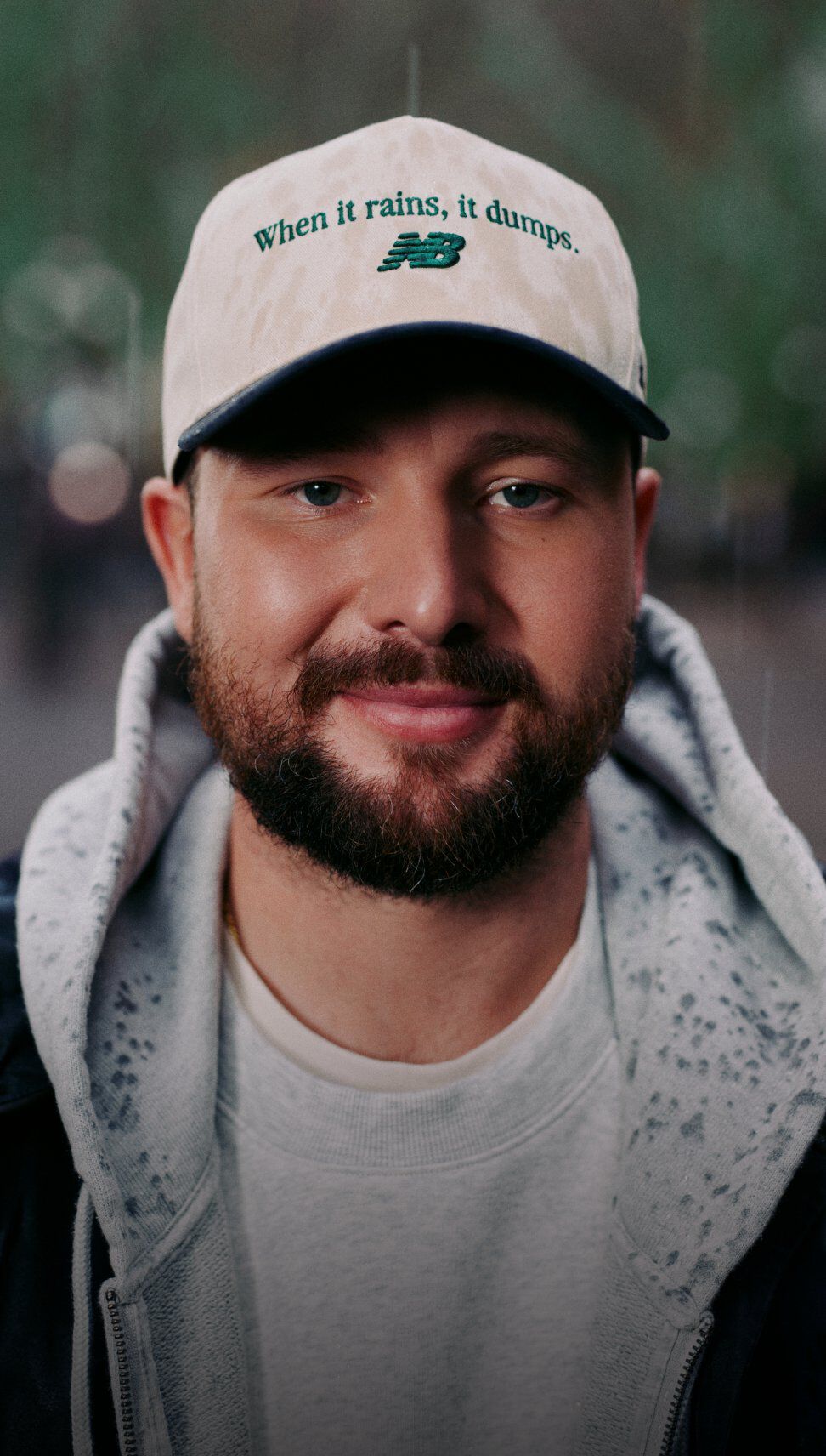Close-up portrait of Cal Raleigh outdoors in light rain, wearing a light-colored baseball cap with the text &ldquo;when it rains, it dumps.&rdquo; and a New Balance logo, wearing a grey hoodie and black jacket.