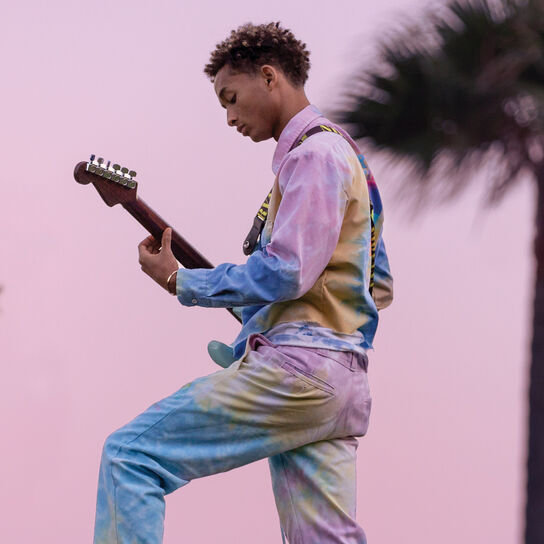 Jaden Smith wearing tie-dye outfit and playing guitar with a pink outdoor background