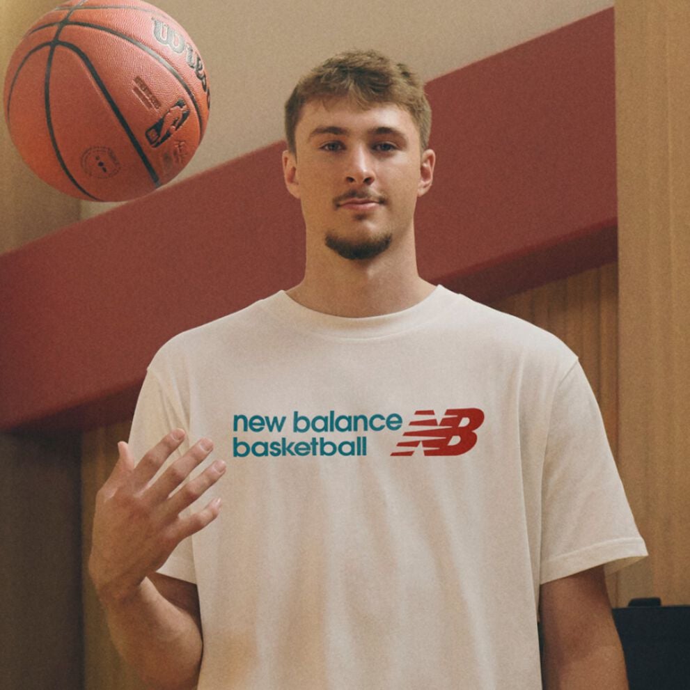 A person wearing a white &ldquo;new balance basketball&rdquo; T‑shirt casually tosses a basketball into the air with one hand while standing indoors against a warm-toned wood and red background.