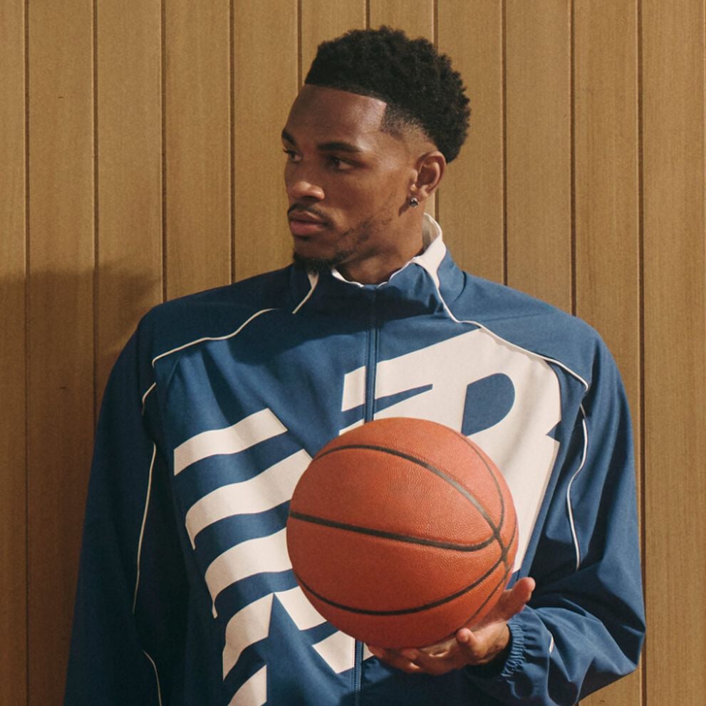 A person wearing a blue New Balance jacket with a large white graphic holds a basketball in front of a wooden-paneled background.