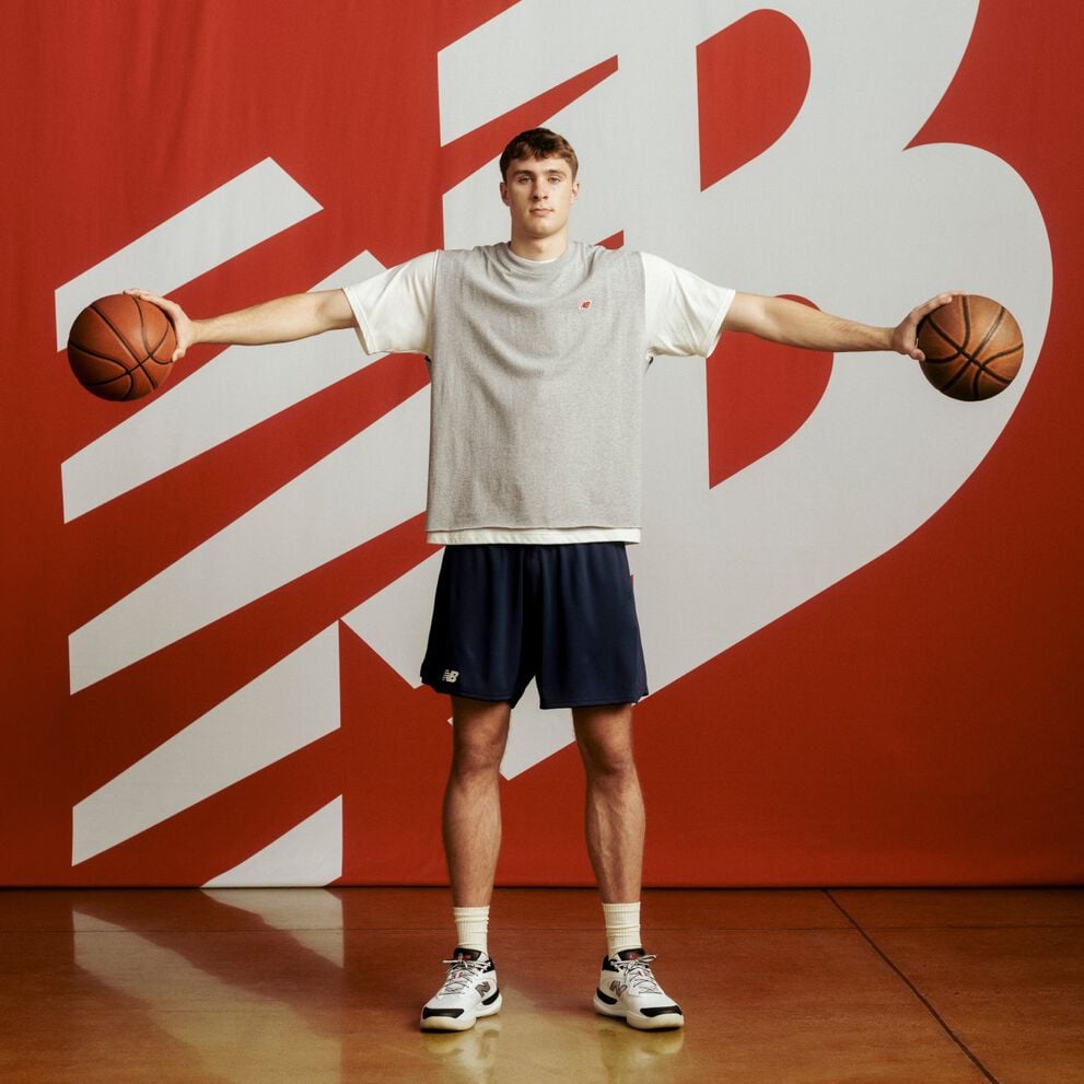 Cooper Flagg de pie frente a un gran fondo rojo con un logotipo blanco de New Balance, vistiendo una camiseta gris sin mangas y pantalones cortos azul marino, sosteniendo una pelota de baloncesto en cada mano con los brazos extendidos horizontalmente.