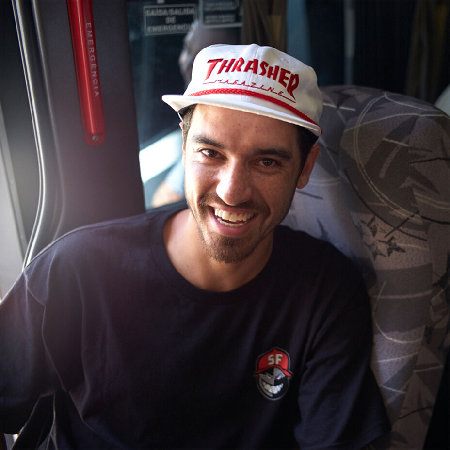 Skateboarder JP Souza in a bus seat, wearing a black graphic tee and white retro snapback