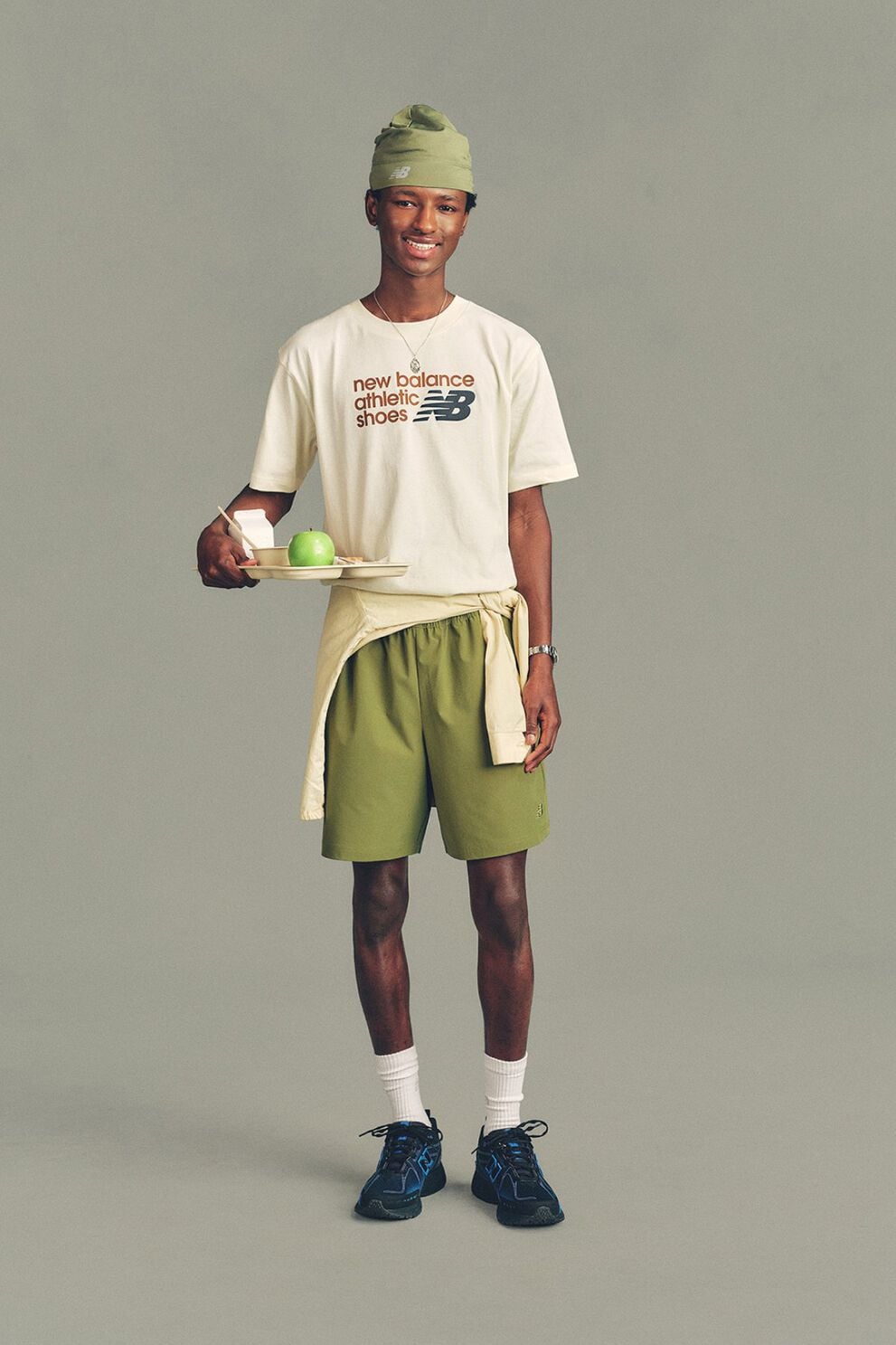 A man in a hat wearing a New Balance shirt and shorts standing in front of a grey studio background.