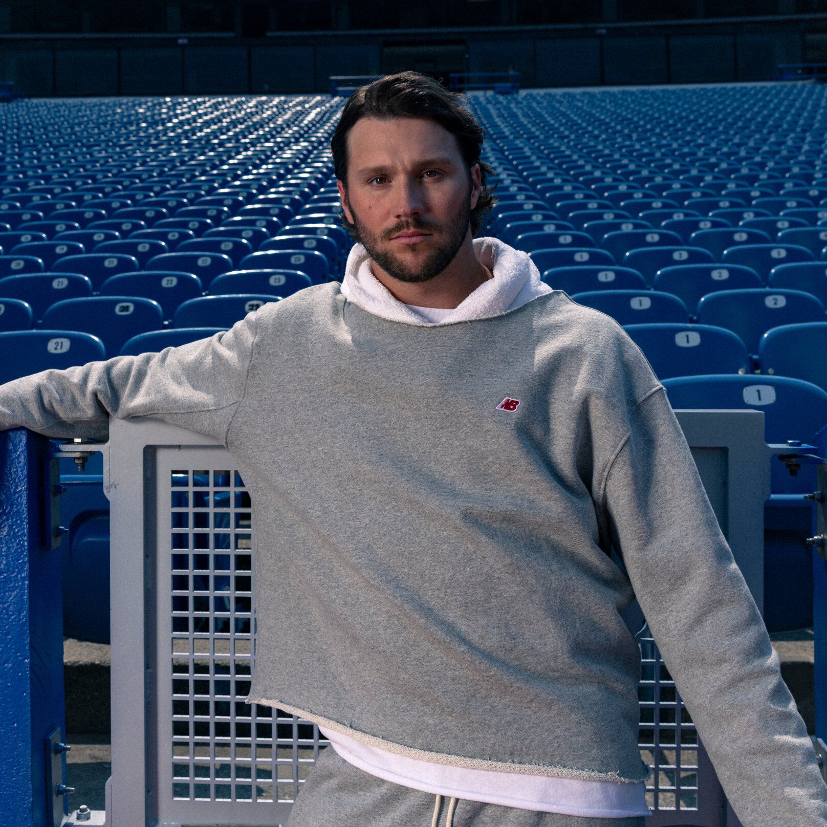 Young male athlete in a grey hoodie leaning on a stadium fence.