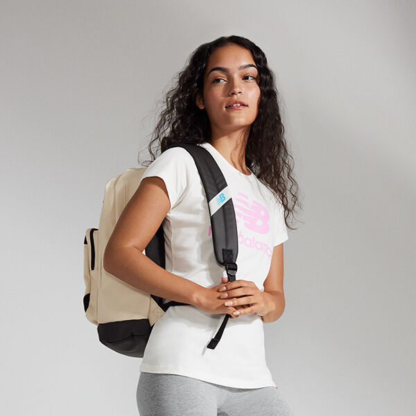 Waist-up image of a young woman wearing a white NB Essentials tee with a pink graphic, grey leggings, and an off-white NB backpack with black shoulder straps 