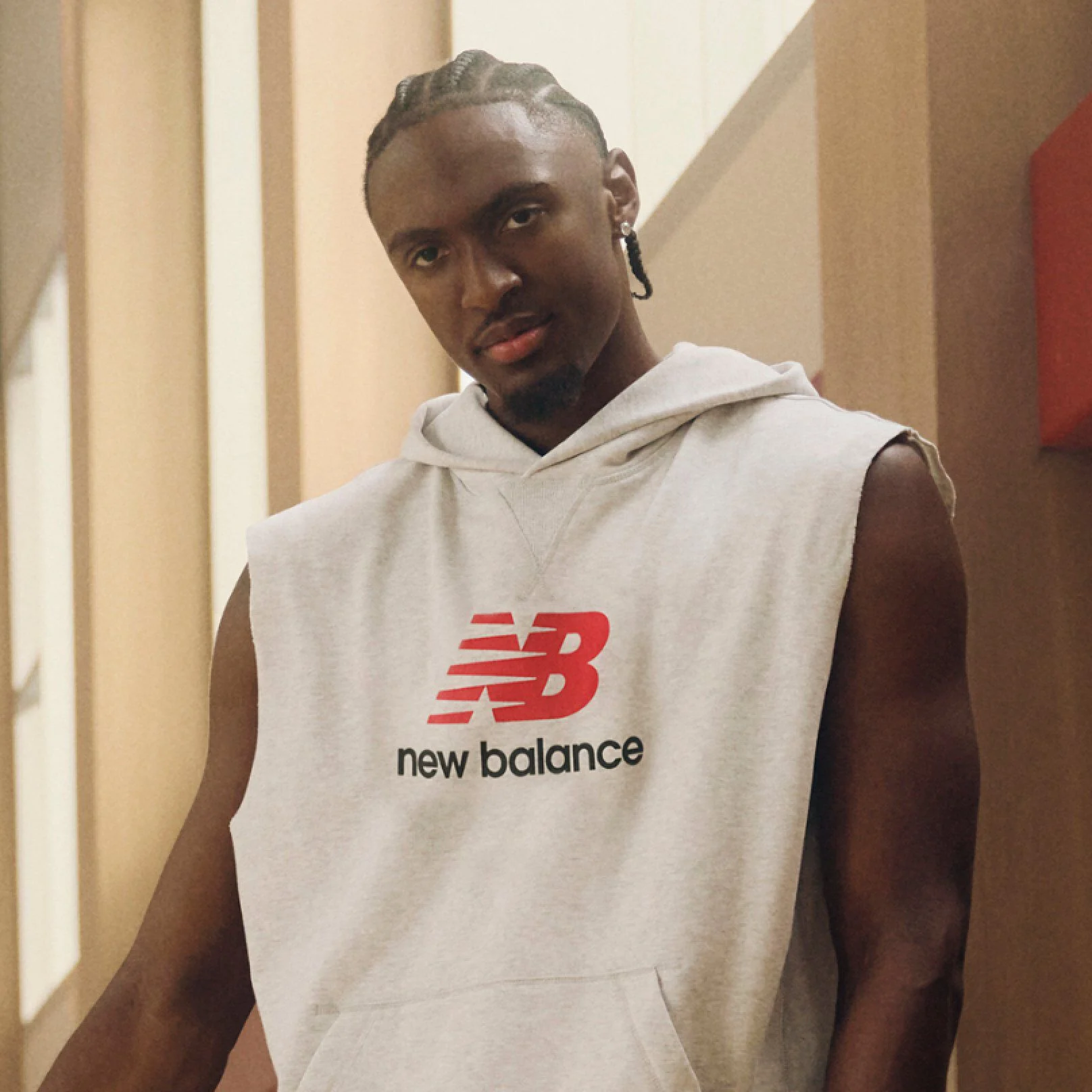 A person stands indoors wearing a sleeveless light‑gray hoodie with a large red New Balance &ldquo;NB&rdquo; logo and the words &ldquo;new balance&rdquo; printed beneath it.