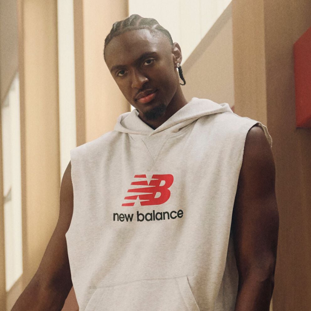 A person stands indoors wearing a sleeveless light‑gray hoodie with a large red New Balance &ldquo;NB&rdquo; logo and the words &ldquo;new balance&rdquo; printed beneath it.