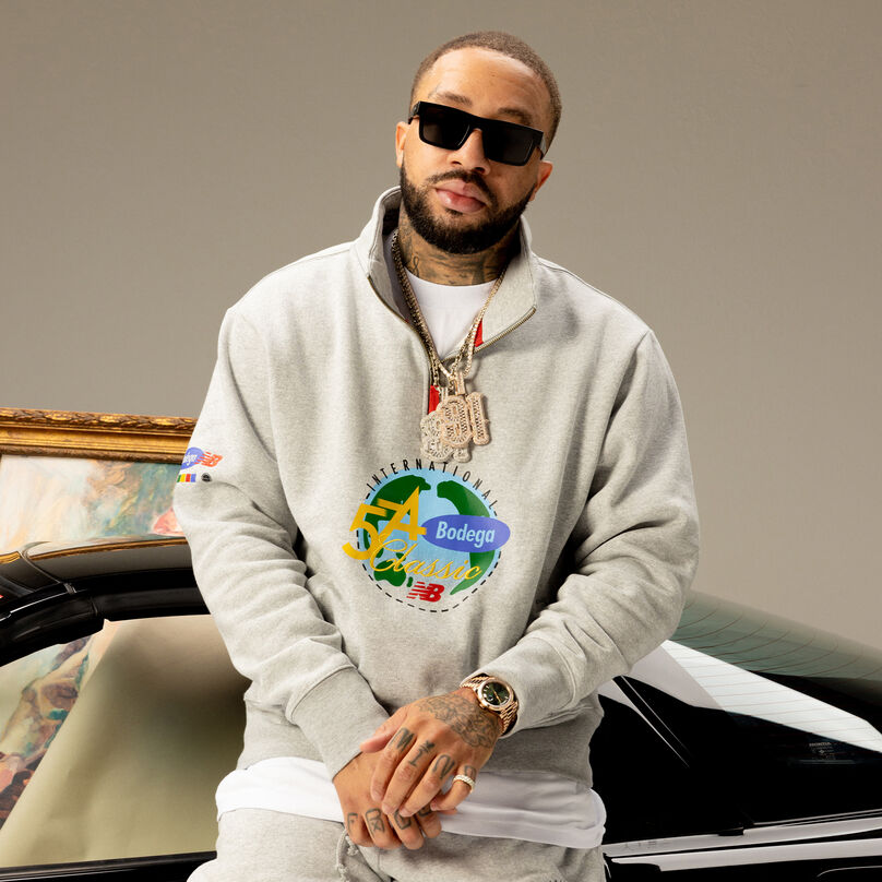Young man in a grey sweatsuit with sunglasses on posing in front of a black car.