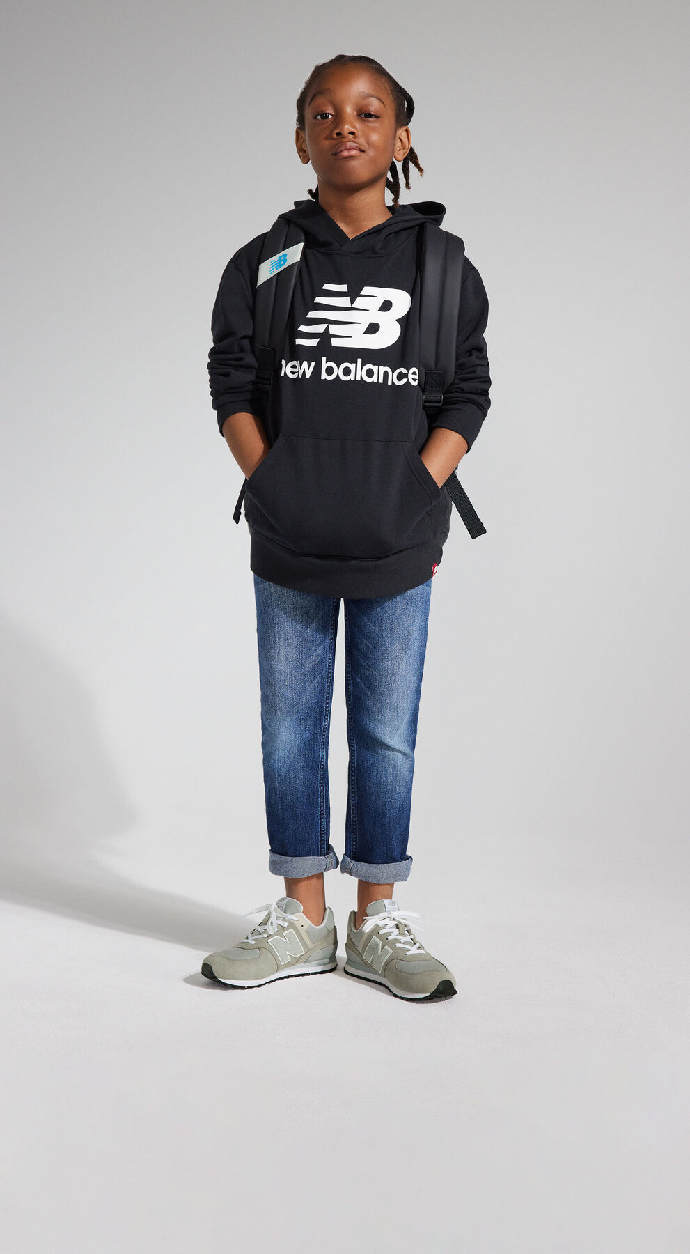 A young boy standing confidently wearing a black NB Essentials hoodie with denim pants and grey NB Classic 574 sneakers in front of a light grey studio backdrop