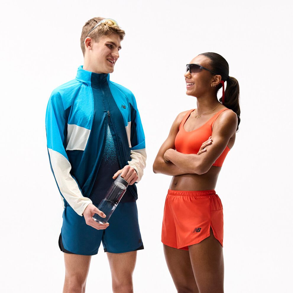 Two people in colorful athletic outfits stand facing each other on a plain white background, one holding a water bottle, both wearing sporty accessories and appearing mid-conversation. 