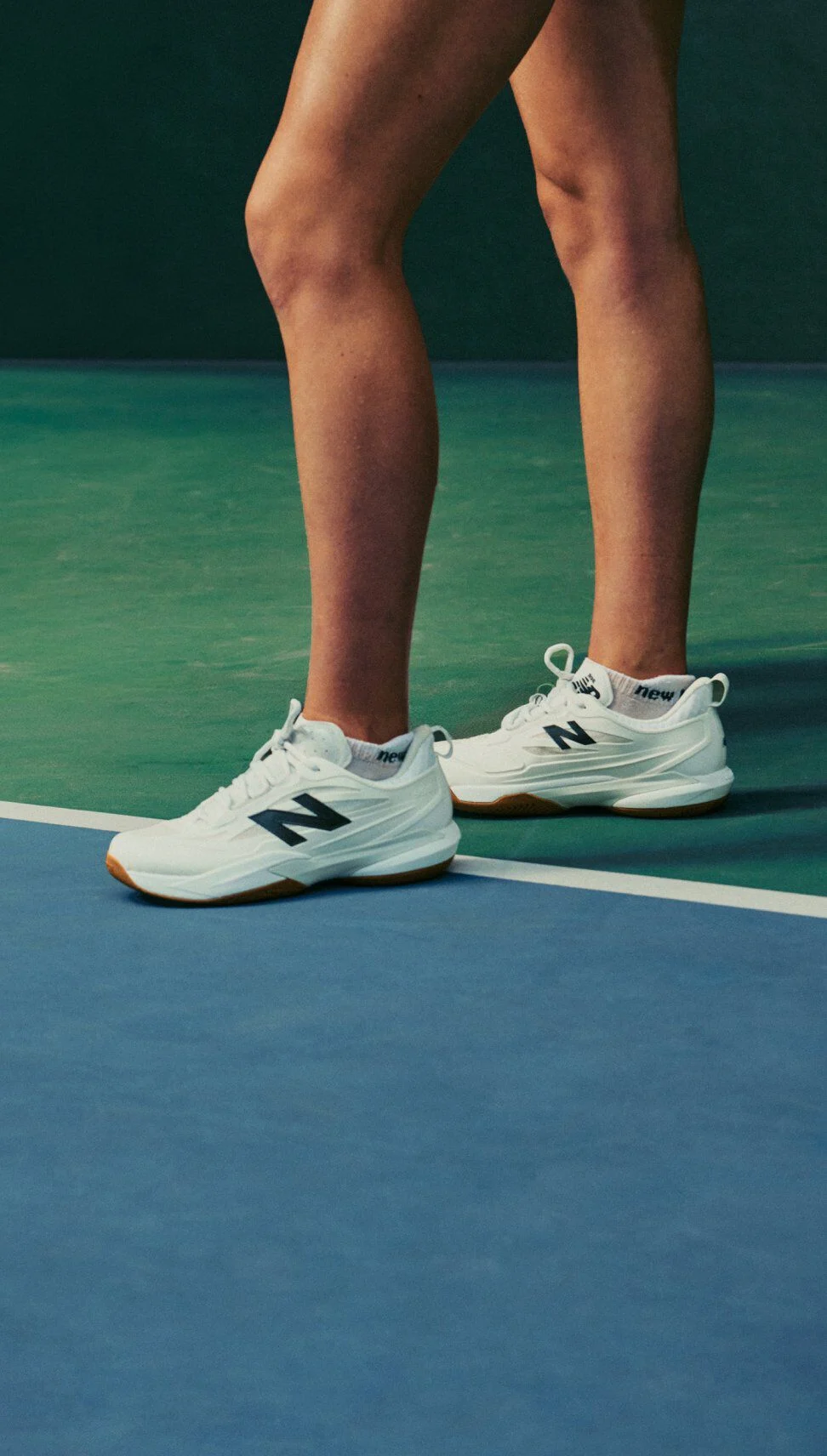 Primer plano de unas zapatillas de tenis New Balance blancas llevadas junto a la pista en una pista cubierta azul y verde.