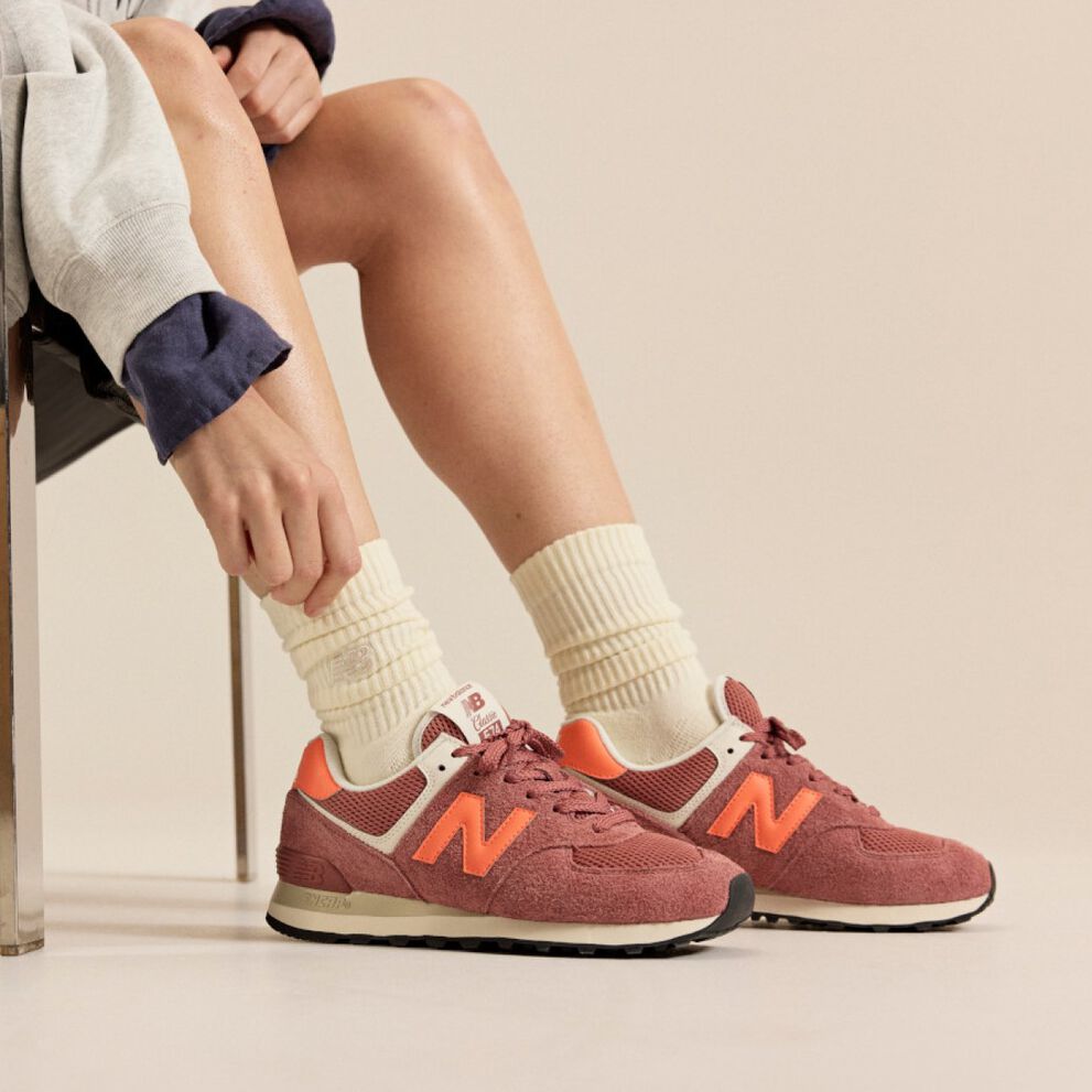 Person sitting on a chair wearing brick‑red New Balance sneakers with orange accents and cream socks against a neutral background. 